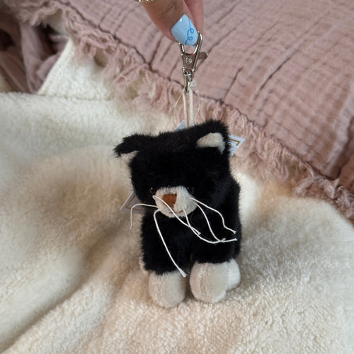 Black and white plush cat with a silver clasp on a soft white surface with a pink textured textile in the background.