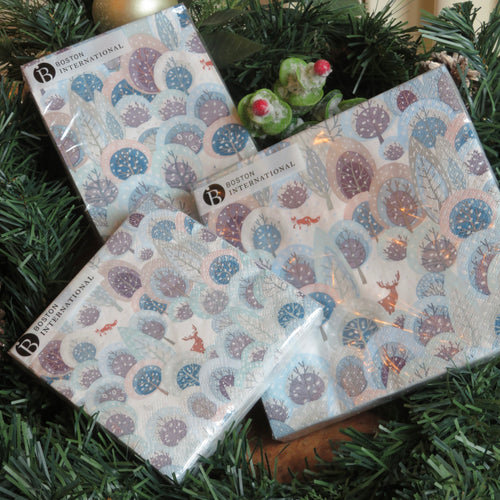 Three packs of paper napkins in assorted sizes- cocktail, guest, lunch- featuring a snowy winter forest design with deer, fox, and stylized trees in shades of blue, lavender, and gray. Displayed among garland and  frosted red berries.  