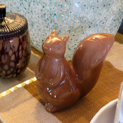 Brown ceramic squirrel figurine on a tan and white textured striped surface with a blue and white speckled object, and a brown and tan container with a brown lid in the background.