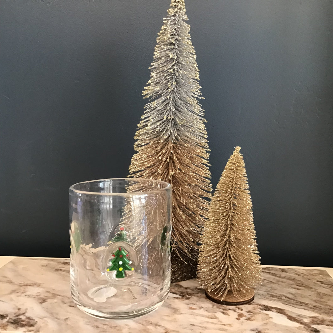A glass tumbler with a green Christmas tree design with red, white, and yellow decorations.  Two decorative frosted Christmas trees behind the glass.  Tumbler and decorative trees on a cream and grey marble slab. Against a blue background. 