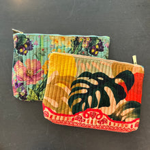 Load image into Gallery viewer, Two quilted velvet zip pouches featuring multicolor designs, including a spring floral pattern in pink, yellow, purple, and green on a light turquoise background and an exotic palm leaf pattern in dark green, tan, yellow, and orange with bottom red border trim, displayed overlapping on a dark surface.
