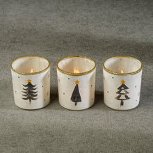 Load image into Gallery viewer, Three white tea light holders with gold glitter rims and simple black Christmas tree designs, each glowing with a lit candle, displayed on a gray textured surface.  