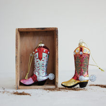 Load image into Gallery viewer, Hand-Painted Cowboy Boot Ornament, two glass cowboy boots featuring a pink toe with a light turquoise shaft and white and red detailing and a gold-colored toe with a red shaft and olive-green detailing, both accented with clear rhinestones, silver-tone spurs, and gold hanging cords, displayed in and beside a rustic wooden box with scattered dirt along the base against a white background. 