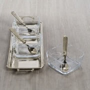 Rectangular metal condiment tray with side handles, holding three square clear glass bowls with matching spoons, next to a separate square glass bowl with a matching spoon, all on a light-colored surface.  