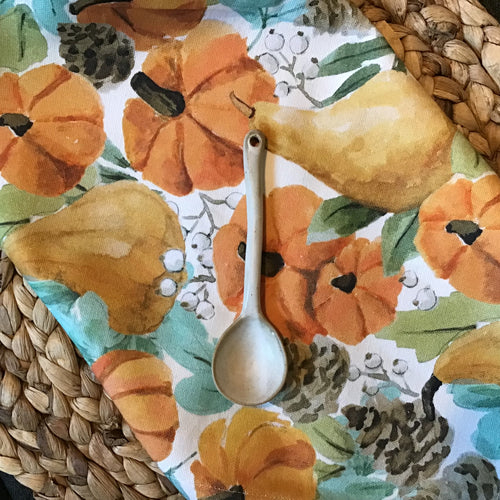 Folded, fabric ,white table runner with an orange pumpkin and gourd design with brown pinecones, green leaves, and white berries. Wooden spoon on runner. Woven surface.