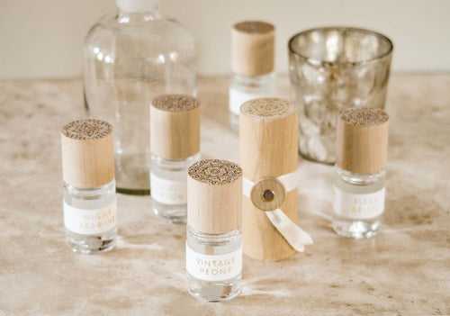 Five roller perfumes with wooden tops on a marble countertop with a glass vase and candle jar. 