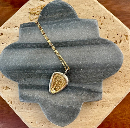 Gold linked chain necklace with a  guardian angel pendant featuring a gold raised design framed by a silver outline, displayed on a quatrefoil-shaped gray stone tile layered over a beige travertine slab on a wooden surface.  