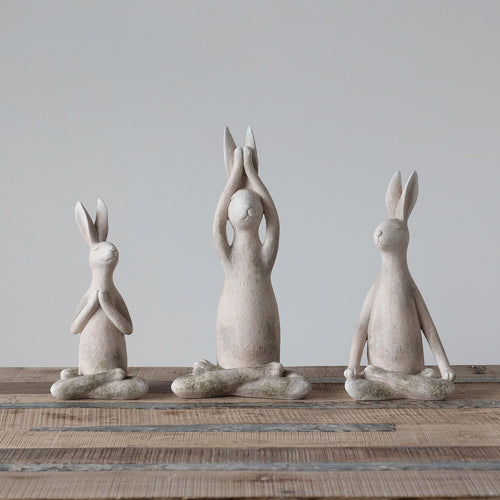 A set of three antiqued white colored resin rabbit figurines in different yoga poses, placed on a wooden surface against a neutral background.