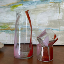 Load image into Gallery viewer, Splash Cup clear hand-blown glass with abstract light pink and red swirled design, stacked on one another beside a matching pitcher on wooden surface against abstract background. 