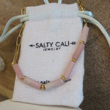 Load image into Gallery viewer, Necklace with pink beads and gold accents on a &quot;Salty Cali Jewelry&quot; pouch on sandy background.