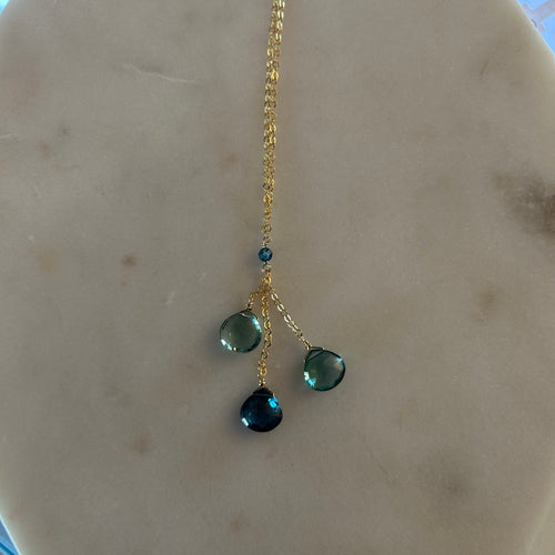 Gold chain necklace with three teardrop gems in various shades of blue. White marble background. 