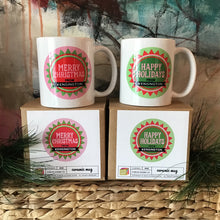 Load image into Gallery viewer, Two white mugs sitting on their cardboard boxes. One mug and box pair is mainly pink and reads &quot;merry christmas from Kensington&quot; and the other is mainly green that reads &quot;happy holidays from Kensington&quot;. Background is an abstract painting and pine needles. 