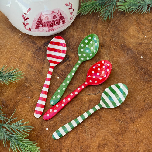 A red and white striped stainless steel spoon. A green stainless steel spoon with white polka dots. A red stainless steel spoon with white polka dots. A green and white striped stainless steel spoon. On a brown wooden surface with green fir tree cuttings , and a white ceramic pot with a red cottage, trees, and botanical motif.