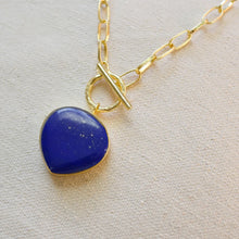 Load image into Gallery viewer, Gold-plated brass chain necklace with a large blue lapis stone pendant and a toggle closure, displayed on a textured cream surface. 