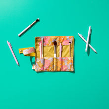 Load image into Gallery viewer, Opened floral makeup brush roll with a mustard yellow interior and pink, orange, lavender, and white retro flower pattern on  the edges, displayed on a teal background with makeup brushes and pens arranged around it. 