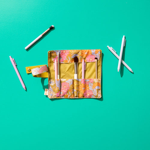 Opened floral makeup brush roll with a mustard yellow interior and pink, orange, lavender, and white retro flower pattern on  the edges, displayed on a teal background with makeup brushes and pens arranged around it. 