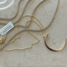 Load image into Gallery viewer, 14K gold-filled necklace featuring a large hammered crescent-moon pendant on a delicate gold chain, displayed on a tan textured surface.