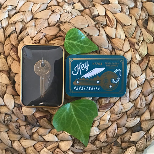 Pocket knife in the shape of a key connected to a keyring. Knife is in a gold tin with the lid sitting next to it. The lid is blue with a picture of the knife printed on it. All sits next to two leaves on a braided brown placemat. 