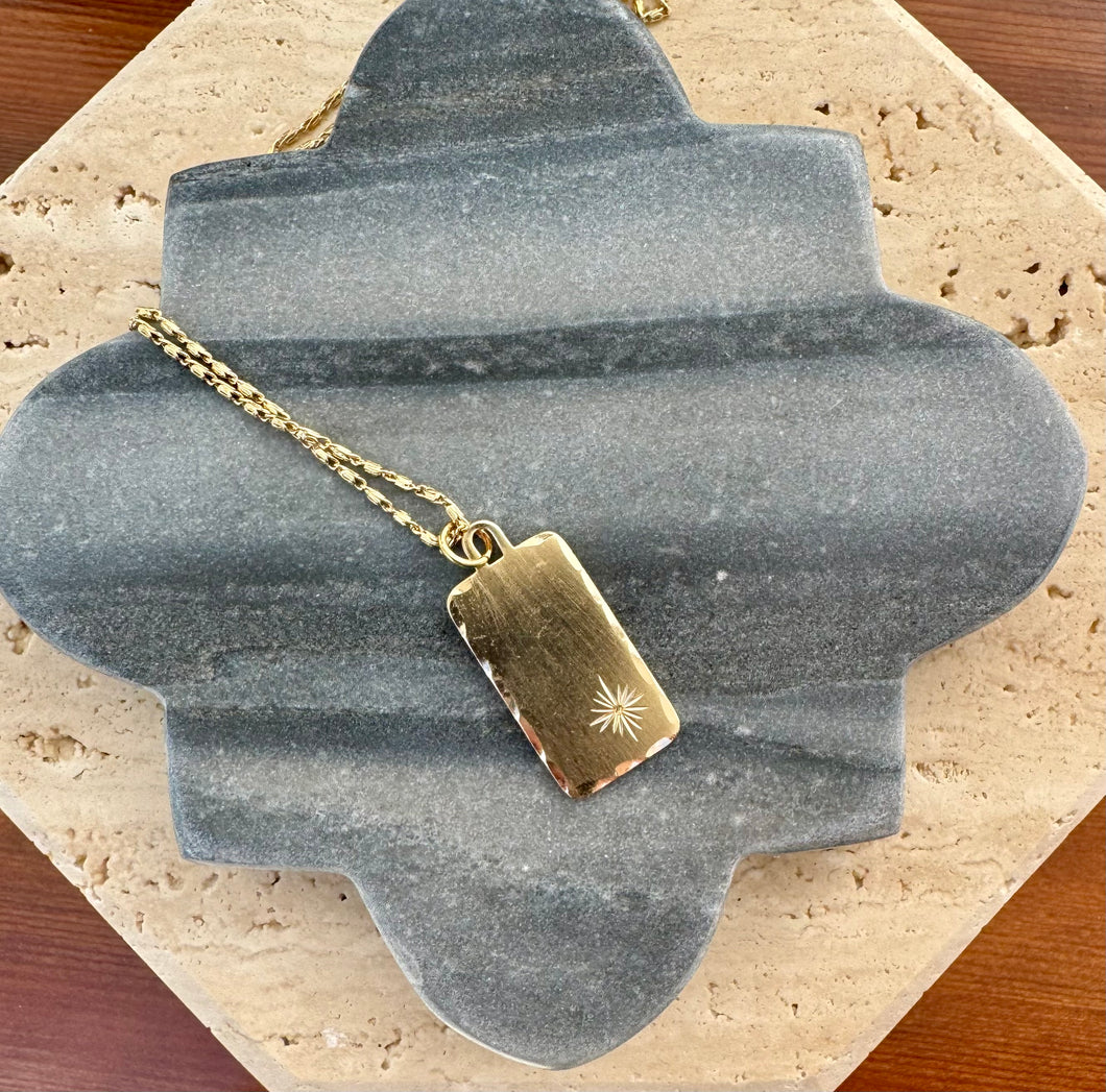 Delicate gold chain necklace with a minimalist flat rectangular pendant featuring an engraved starburst detail, displayed on a quatrefoil-shaped gray stone tile layered over a beige travertine slab on a wooden surface.  