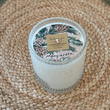 Load image into Gallery viewer, Balsam &amp; Fir, lifestyle image showing an angled overhead view of a cream soy candle in a clear glass jar with raised dot texture and an unlit single wick, topped with decorative paper featuring green evergreen branches, brown pinecones, and a gold-colored panel with the scent name and product type in dark lettering, resting on a brown woven mat.
