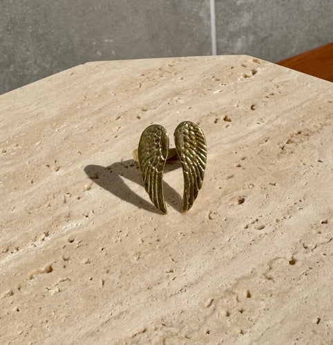 Gold-tone ring featuring two detailed angel wings, displayed on a beige travertine slab.