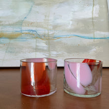 Load image into Gallery viewer, Splash Cup clear hand-blown glass set of two with abstract light pink and red swirled design on wooden surface against abstract background.