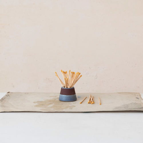 A stoneware match holder with striker on a wooden surface, filled with matches, against a neutral background.