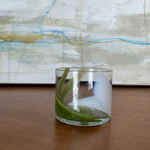 Load image into Gallery viewer, Splash Cup clear hand-blown glass with abstract pistachio green and white swirled design on wooden surface against abstract background.