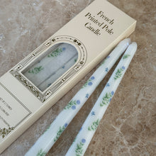 Load image into Gallery viewer, Two white taper candles with blue-and-green floral designs on a brown marble surface, next to their packaging.