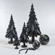 Matte black decorative trees in varying heights with layered, angular branches, displayed with two round matte black decorative balls featuring a glitter gold leaf-like designs, one small with a black ribbon and one larger, on a white surface. 