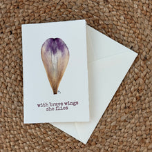 Load image into Gallery viewer, Colby Cards, lifestyle image of a white Lokta paper greeting card featuring a real pressed purple and cream flower petal above “With Brave Wings She Flies” dark purple lettering, displayed with a white envelope on a brown woven mat.