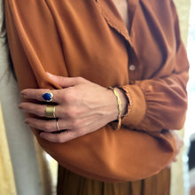 Load image into Gallery viewer, Model wearing a double-gold-plated brass ring with an oval blue lapis stone and CZ stone accents, styled with coordinating gold rings and gold bracelet, shown against a warm tan blouse.   