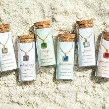 Load image into Gallery viewer, Six 14K gold-dipped birthstone bottle necklaces displayed in clear glass jars with cork lids on a sandy surface, each featuring a square glass crystal pendant representing different birth months. 