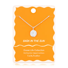 Load image into Gallery viewer, Sterling silver-dipped necklace featuring a small sun charm pendant and a chain with delicate beaded accents, displayed on a golden yellow &quot;Bask in the Sun&quot; Ocean Life Collection card by Lucky Feather with a white wavy border against white background. 