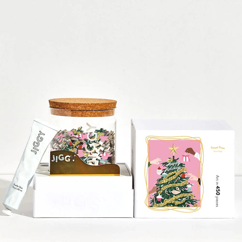 White opened box with a glass jar and a cork lid with a jigsaw puzzle taken apart inside. On the box top shows the puzzle print which is a Christmas tree being decorated on a pink background. A tube of puzzle glue is propped up on the left. All on a white background. 