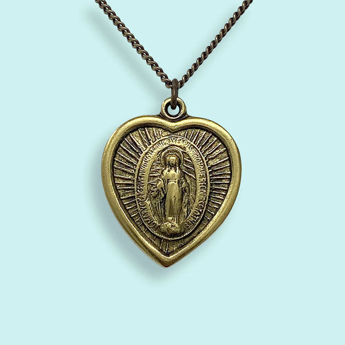 Heat pendant stamped with the shape of Mother Mary on a gold chain. Light blue background. 