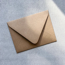 Load image into Gallery viewer, Brown kraft paper envelope shown enclosure flap side up on a light gray textured fabric surface.