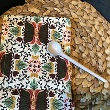 Load image into Gallery viewer, Cotton kitchen towel in cream with a floral and leaf pattern in green, gold, and burgundy, displayed on a brown woven surface with a white long handle spoon resting on top. 