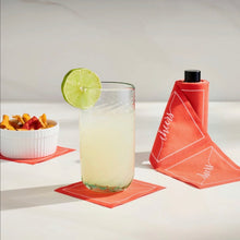 Load image into Gallery viewer, Matte black cocktail napkin stand holding a roll of red cocktail napkins with "cheers" in white script, displayed on a white surface beside a drink with a lime garnish and a white snack bowl filled with snacks, both resting on red cocktail napkins, against a gray wall.