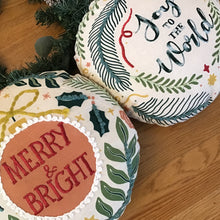 Load image into Gallery viewer, Two white round ornament shaped Christmas pillows side by side on a wooden surface with green garland in the background. First pillow has an orange round center with a white looping edging and a yellow bow tie surrounded by green holly leaves and other green leaves. &quot;Merry &amp; Bright&quot; embroidered in red lettering. The second pillow is white with various green leaves and red and yellow designs. &quot;Joy To The World&quot; in green embroidered letters.   