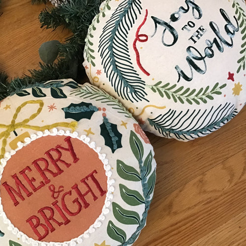Two white round ornament shaped Christmas pillows side by side on a wooden surface with green garland in the background. First pillow has an orange round center with a white looping edging and a yellow bow tie surrounded by green holly leaves and other green leaves. 