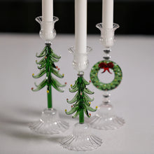 Load image into Gallery viewer, Three glass taper candle holders in festive holiday designs, including a large shaped green Christmas tree, a small shaped green Christmas tree, and one shaped liked a green wreath with a red bow, displayed together on a white surface with white taper candles. 