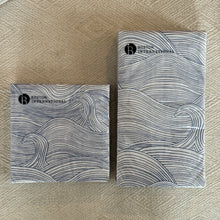 Load image into Gallery viewer, Paper cocktail napkins and guest napkins with blue wave pattern on white background, displayed on woven neutral surface. 