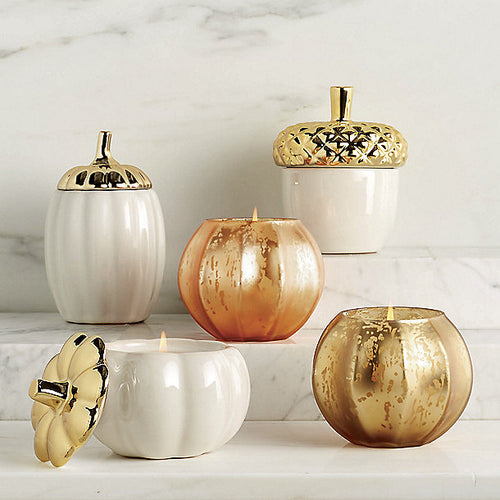 two White ceramic pumpkins and one acorn with gold lids arranged  on marble platforms with a two peach and gold pumpkin candles. White marble background. 