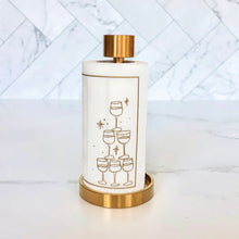 Load image into Gallery viewer, Roll of white cotton cocktail napkins with a gold border and gold outlined stack-of-glasses illustration displayed on a gold napkin stand against a white tile background. 