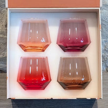 Load image into Gallery viewer, Set of four hand-blown crystal stemless glasses in gradient amber, rose, red, and plum hues, neatly arranged inside a cream-colored gift box with a coral-colored lid, displayed on a wooden surface. 