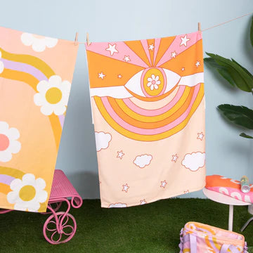 Two colorful retro-style beach towels hanging on a clothesline against a light blue background with artificial green grass below. The towel on the right features an eye and a rainbow design in shades of pink and orange with white stars and clouds. The towel on the left shows large white daisies and curved orange and lavender stripes. A tall green plant, a pink and orange cooler bag , a white table with a folded pink towel, and a partial view of a pink lawn chair in the background. 