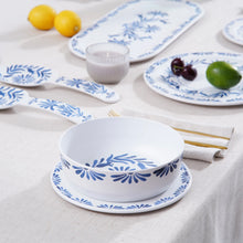 Load image into Gallery viewer, White melamine dinner bowl with a blue floral pattern on a white surface styled with a matching serving fork and spoon, serving platter, plates, candle, and food. 