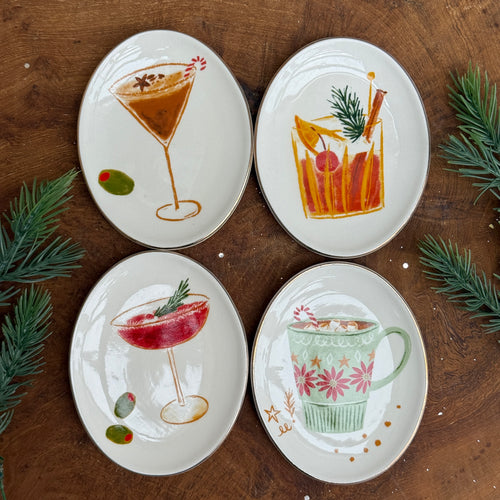 Four white oval ceramic dishes with holiday drink illustrations: a martini glass with a brown drink, candy cane, star garnish, and olive; an orange drink with an orange slice, cinnamon stick, cherry, and fir sprig; a red cocktail with fir garnish and two olives; and a green floral mug with a brown drink, marshmallows, and candy cane. All on a wooden surface with greenery.