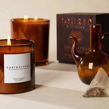 Load image into Gallery viewer, Poured NY Curiositeas Candle | Two Styles, candle with white wax and a single lit wick in a cylindrical translucent dark-amber brown glass vessel beside a taller two-wick lit candle, dark-amber glass teapot, pyramid tea sachet, and a dark Poured New York box against a cream background. 